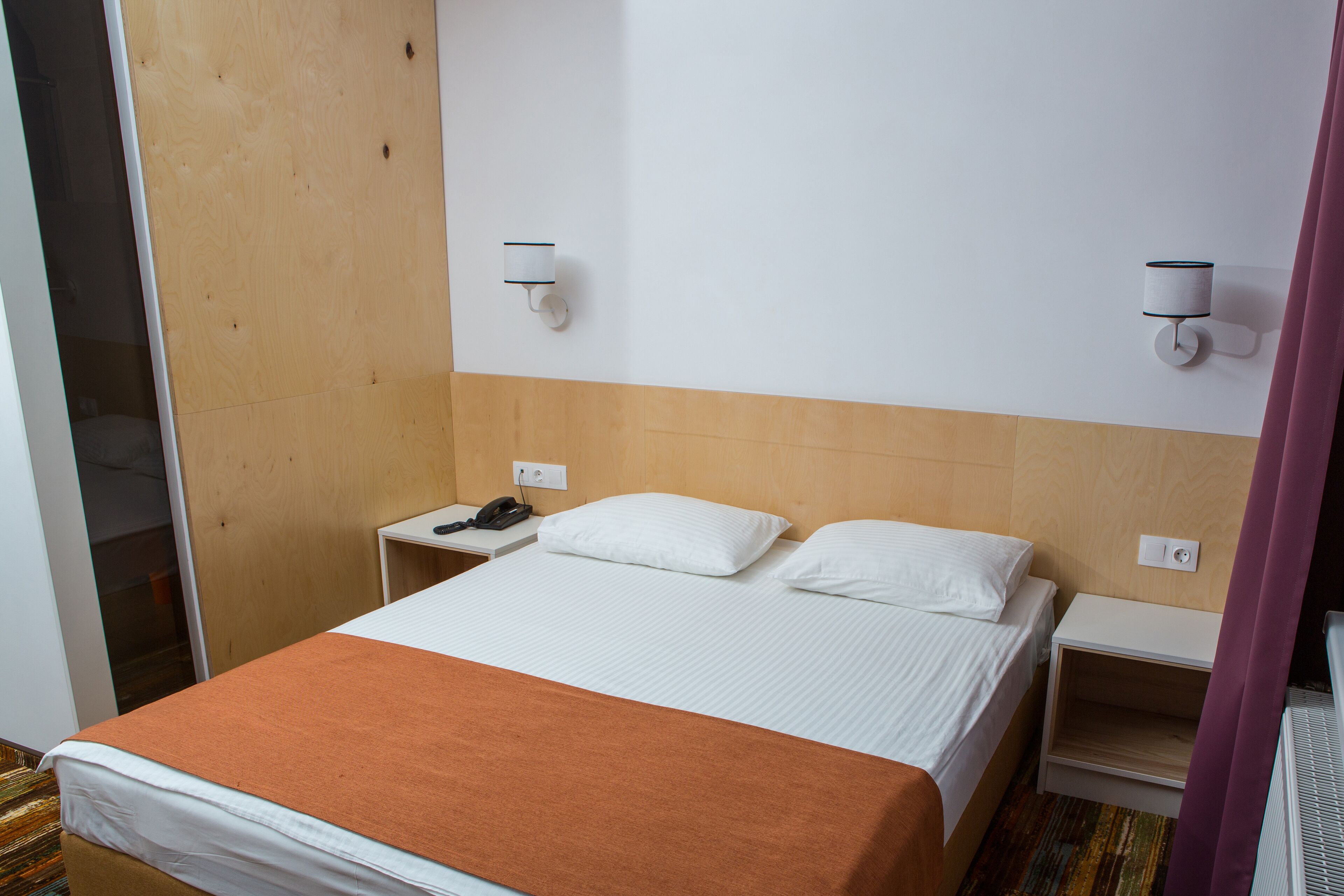 A hotel room with a bed, a nightstand, a telephone, and two wall-mounted lamps.