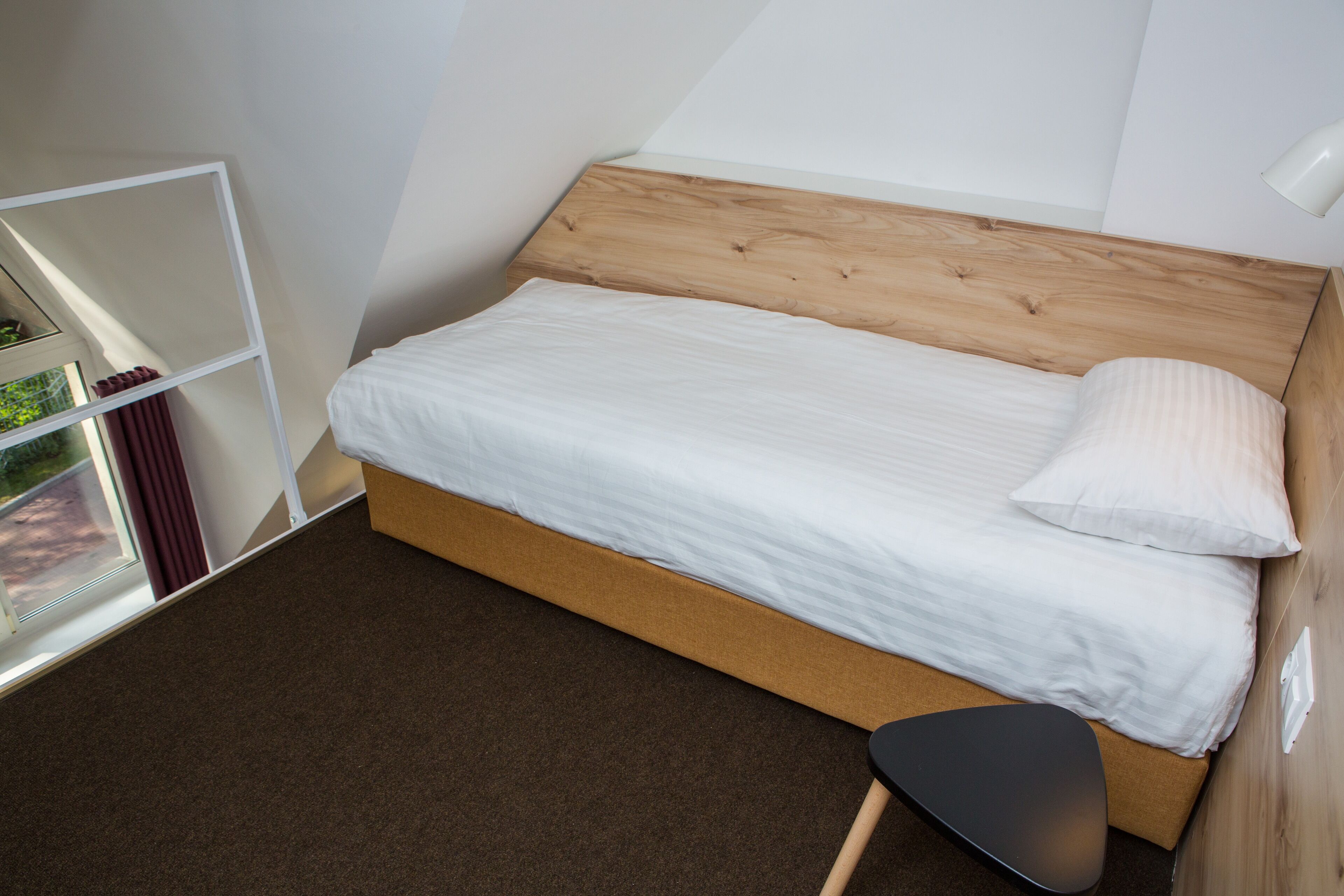 A bedroom with a wooden headboard, a single bed with white bedding, a brown carpet, and a small black stool.