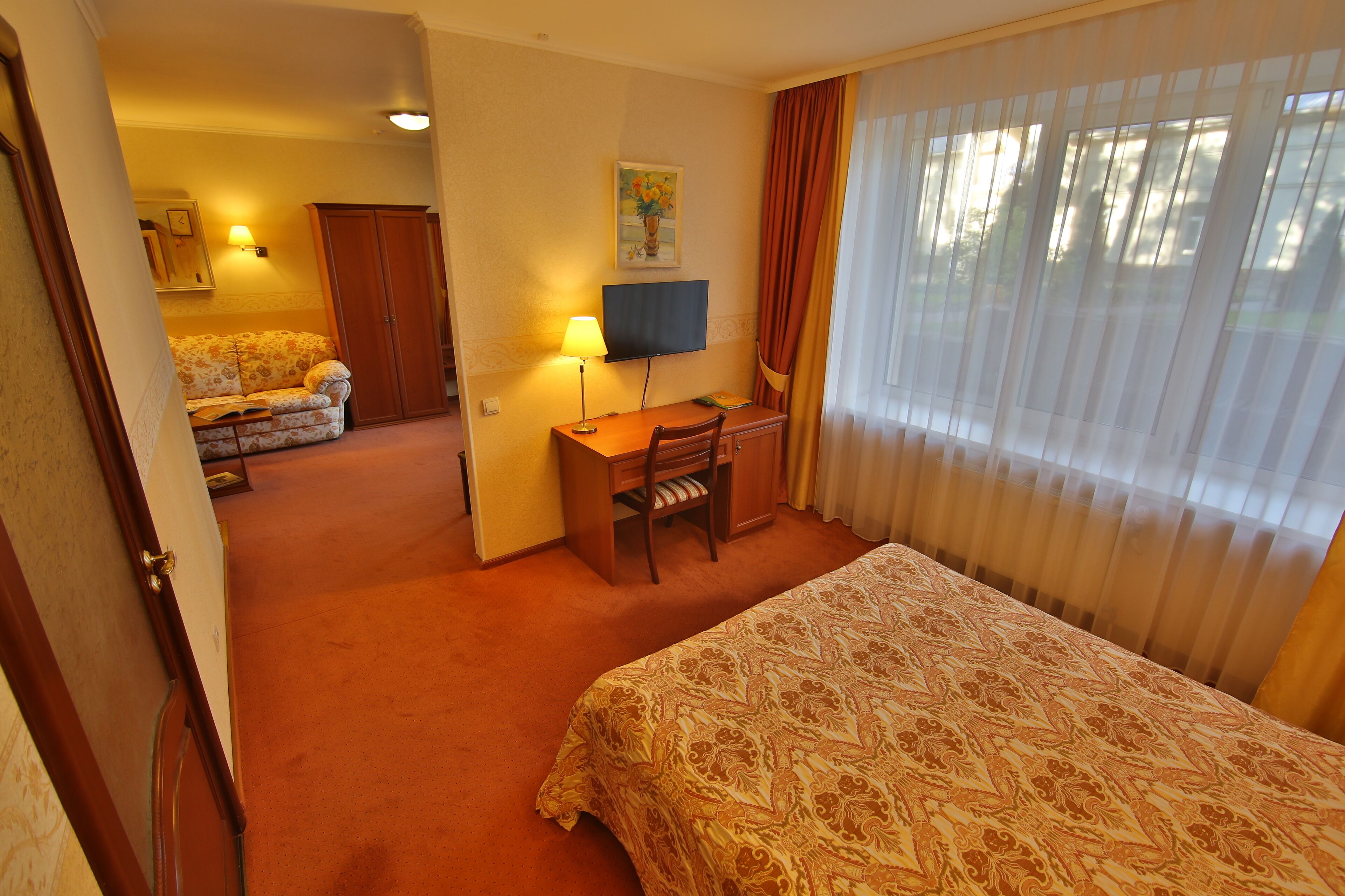 A hotel room with a bed, a desk with a chair, a television, a sofa, and a window with curtains.