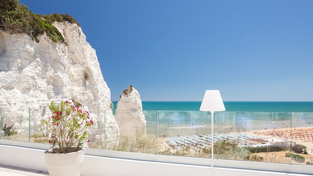 A balcony with a potted plant, a lamp, and a view of a beach with umbrellas and a clear sky. - Hotel Falcone (Vieste)