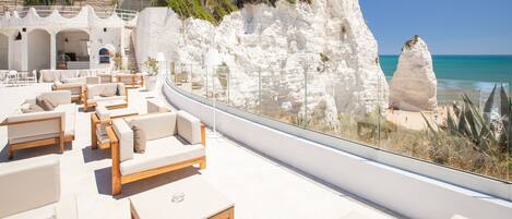 A seaside terrace with white stone walls, cushioned seating, and a clear view of the ocean and cliffs.