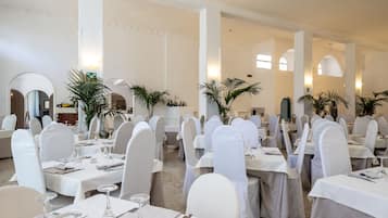 Un comedor amplio con mesas y sillas blancas, dispuesto en una distribución simétrica, con ventanas amplias y arcos decorativos.