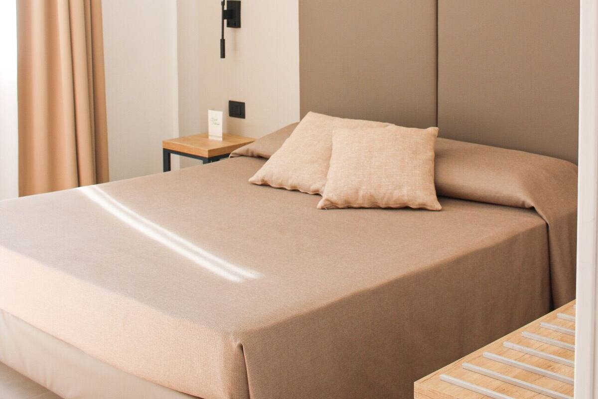 Habitación de hotel minimalista con una cama grande, una mesita de noche, una lámpara y vistas al exterior a través de una ventana con cortinas.