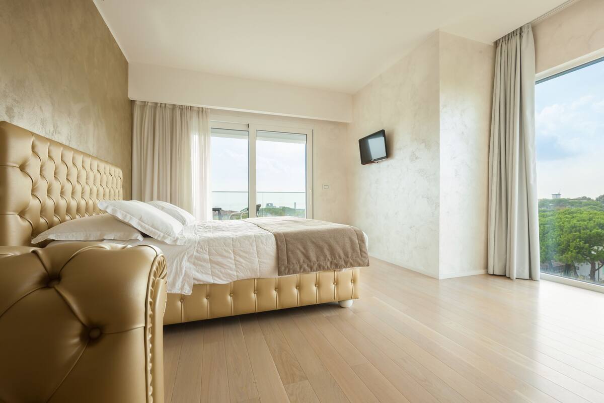 A spacious bedroom with a large bed, a television mounted on the wall, and a view of the outdoors through a large window.