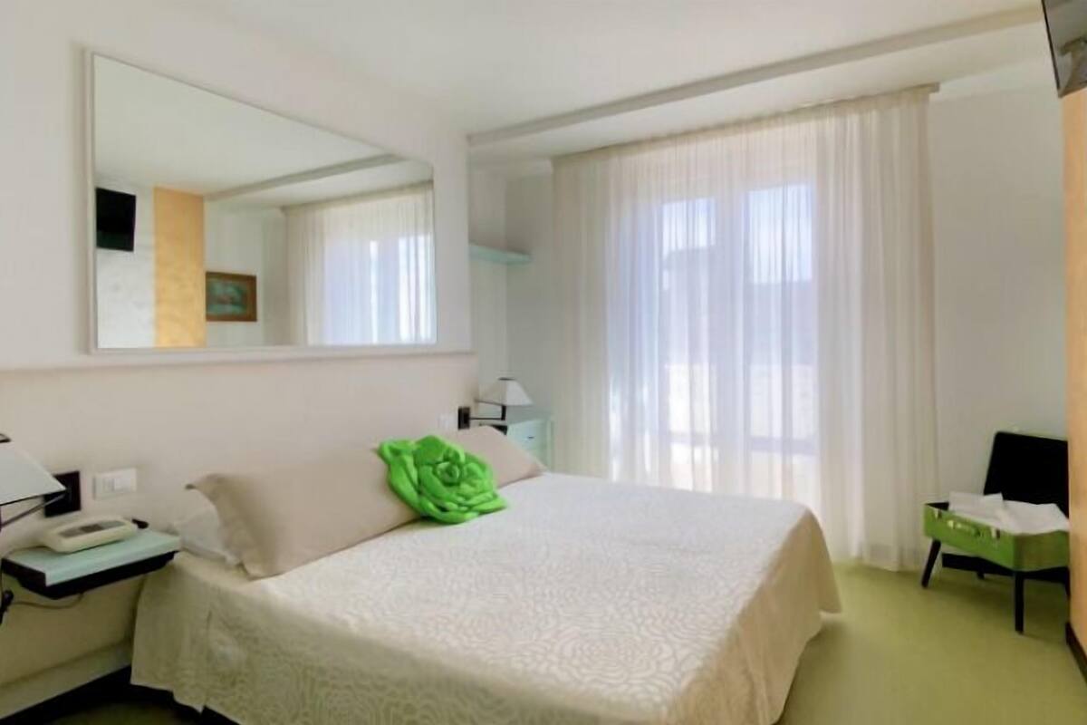 A hotel room with a bed, a bedside table with a lamp, a chair, and a large window with curtains.