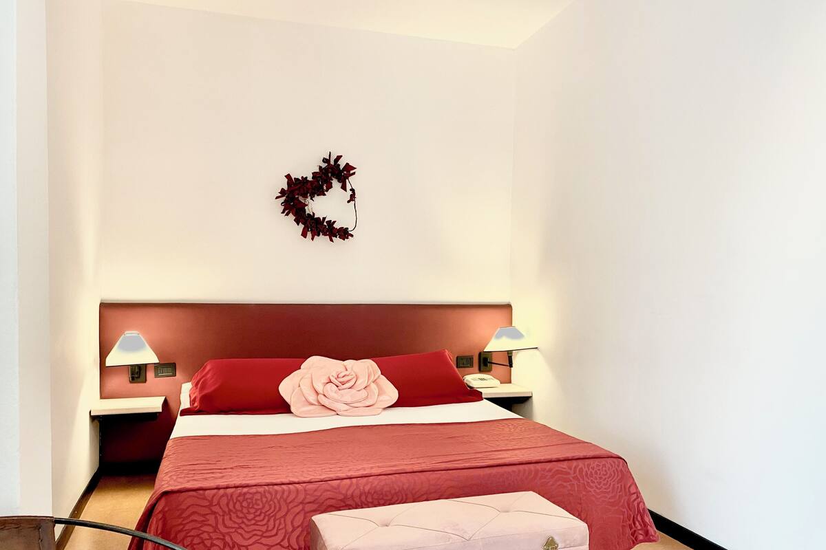 A bedroom with a large bed, a nightstand, a bedside lamp, a decorative wall piece, and a small bench.