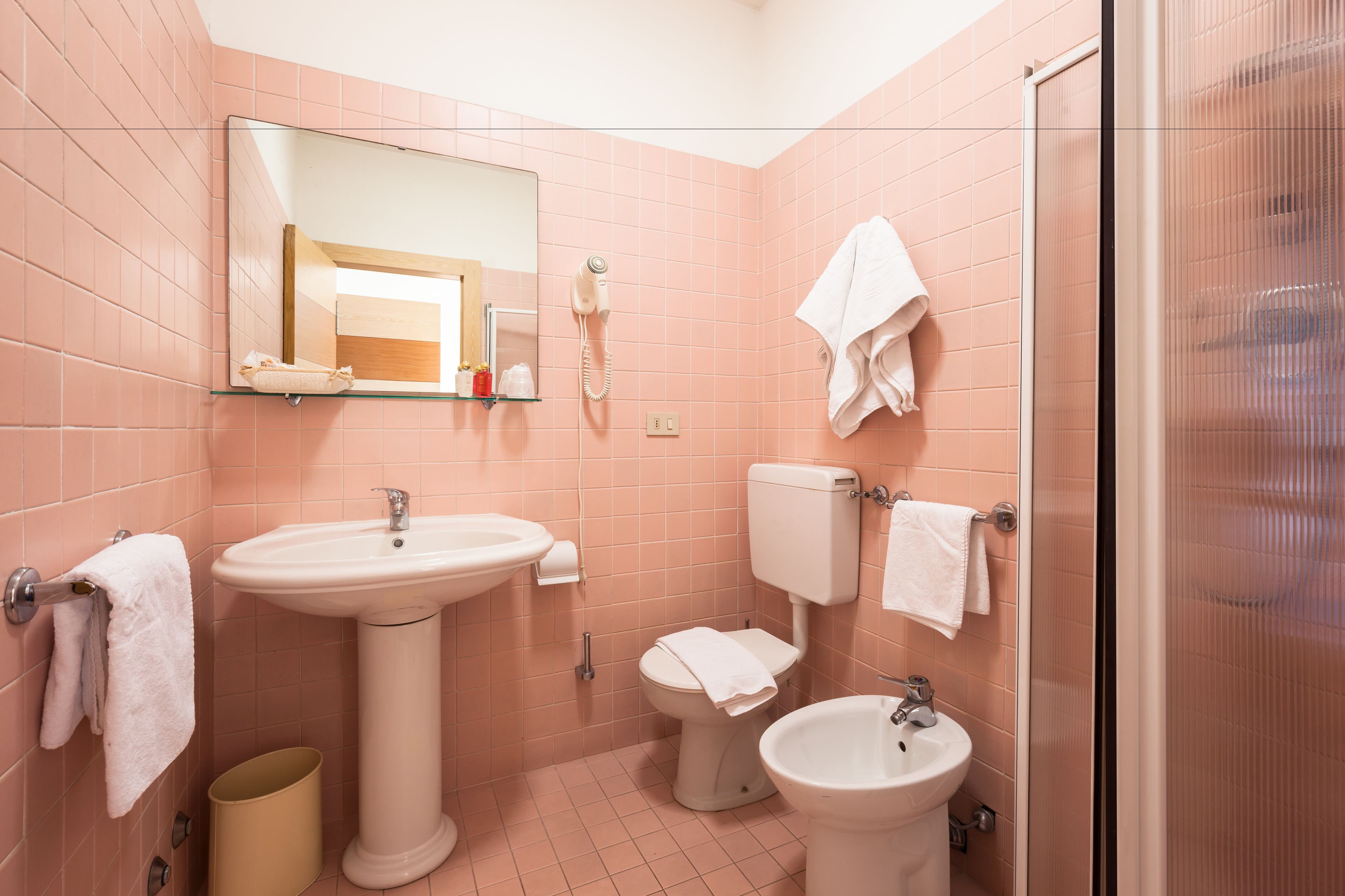 A bathroom with pink tiles, a sink, a toilet, a bidet, a mirror, and a glass shower enclosure.