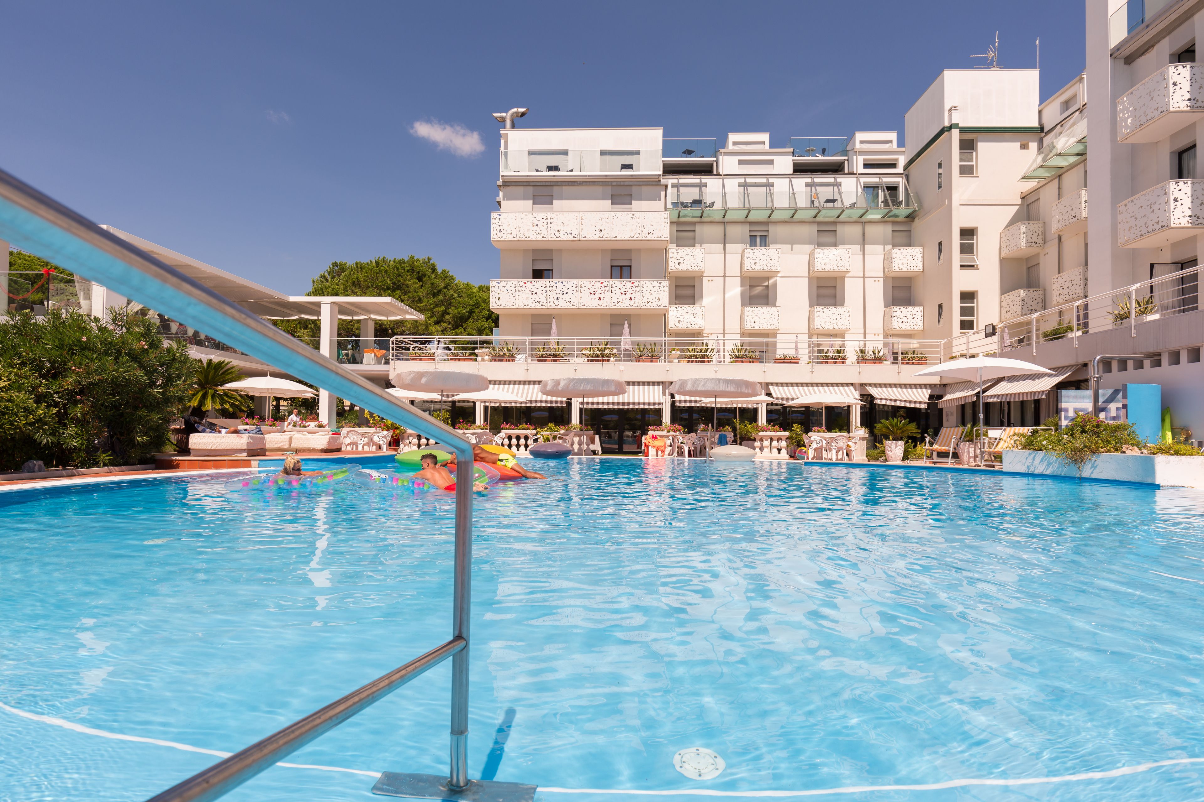 A swimming pool with a clear blue water surface, surrounded by lounge chairs and umbrellas, with a multi-story building in the background.