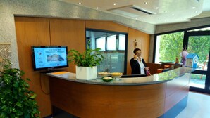 A reception desk with a television displaying an event, a potted plant, and a view of the outdoors. - Centro Vacanze Cavallino Opera Nascimbeni (Cavallino-Treporti)