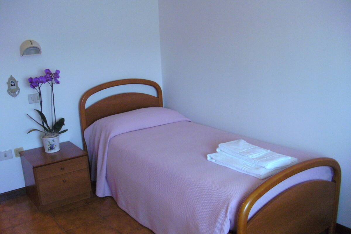 Ein Einzelbett mit Holzrahmen und einer rosa Tagesdecke, ein kleiner Nachttisch mit Pflanze und eine wandmontierte Leuchte.