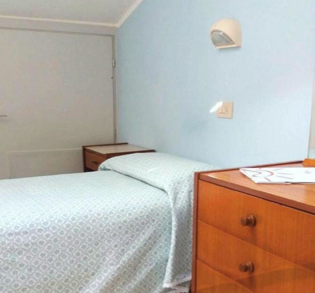 Um quarto pequeno e simples com uma cama, uma mesinha de cabeceira de madeira e uma escrivaninha com uma cadeira.