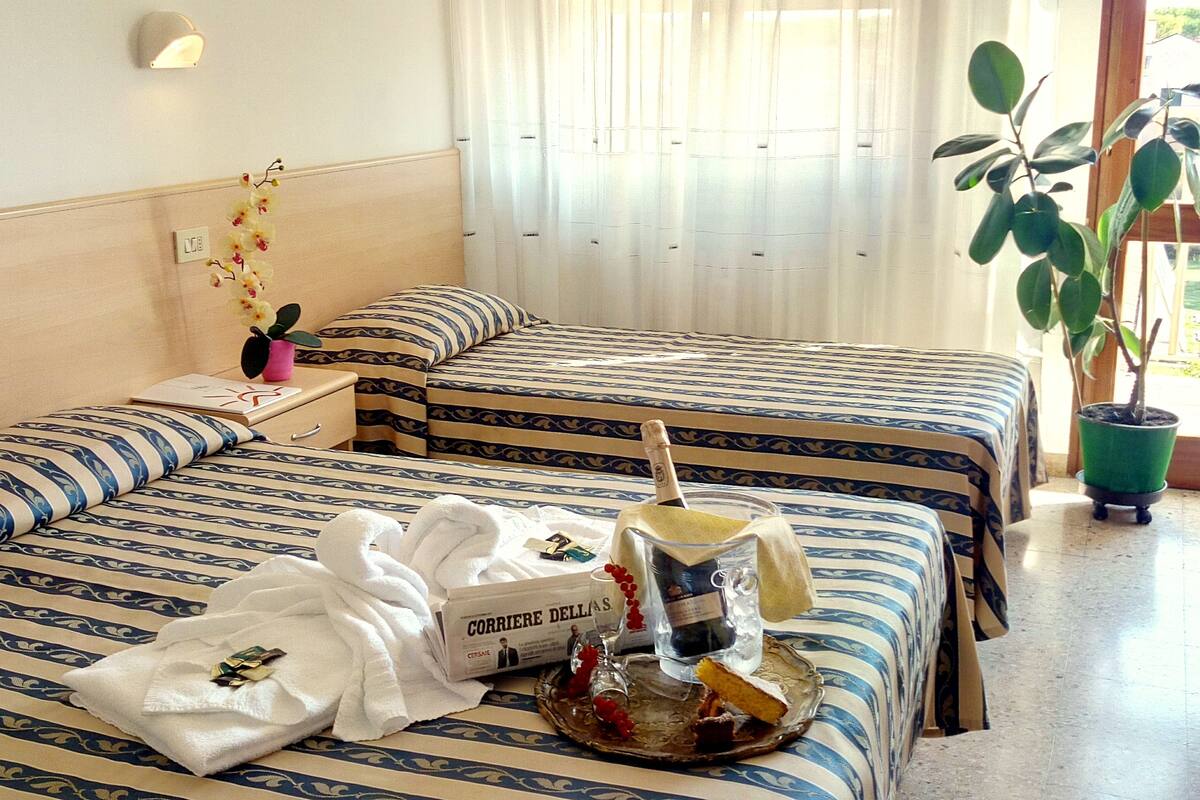 Ein Hotelzimmer mit zwei Betten, einem Nachttisch, einer Topfpflanze und einem Frühstückswagen mit einer Champagnerflasche und einer Schokoladenschachtel.