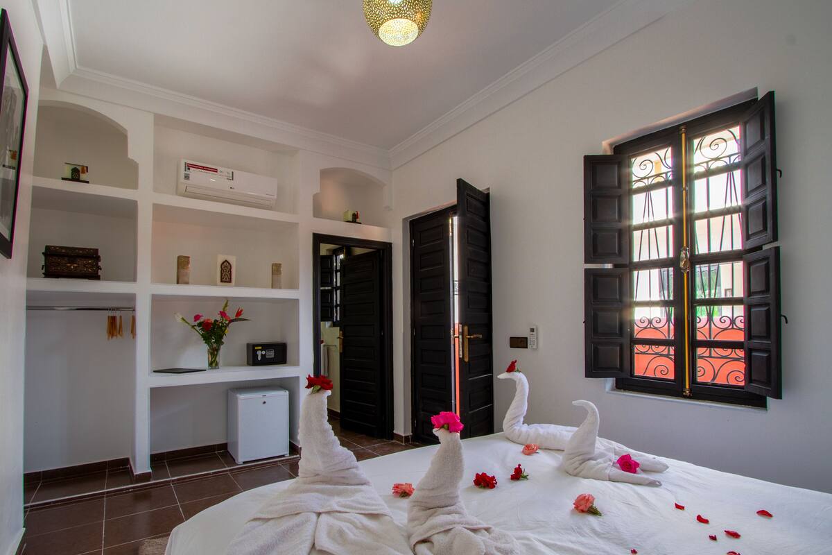A hotel room with a bed, a desk, a chair, a small table, a shelf with decorative items, a window with black shutters, and a door.