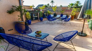 Terraza en la azotea con sillas azules, una mesa con fruta y plantas en macetas.