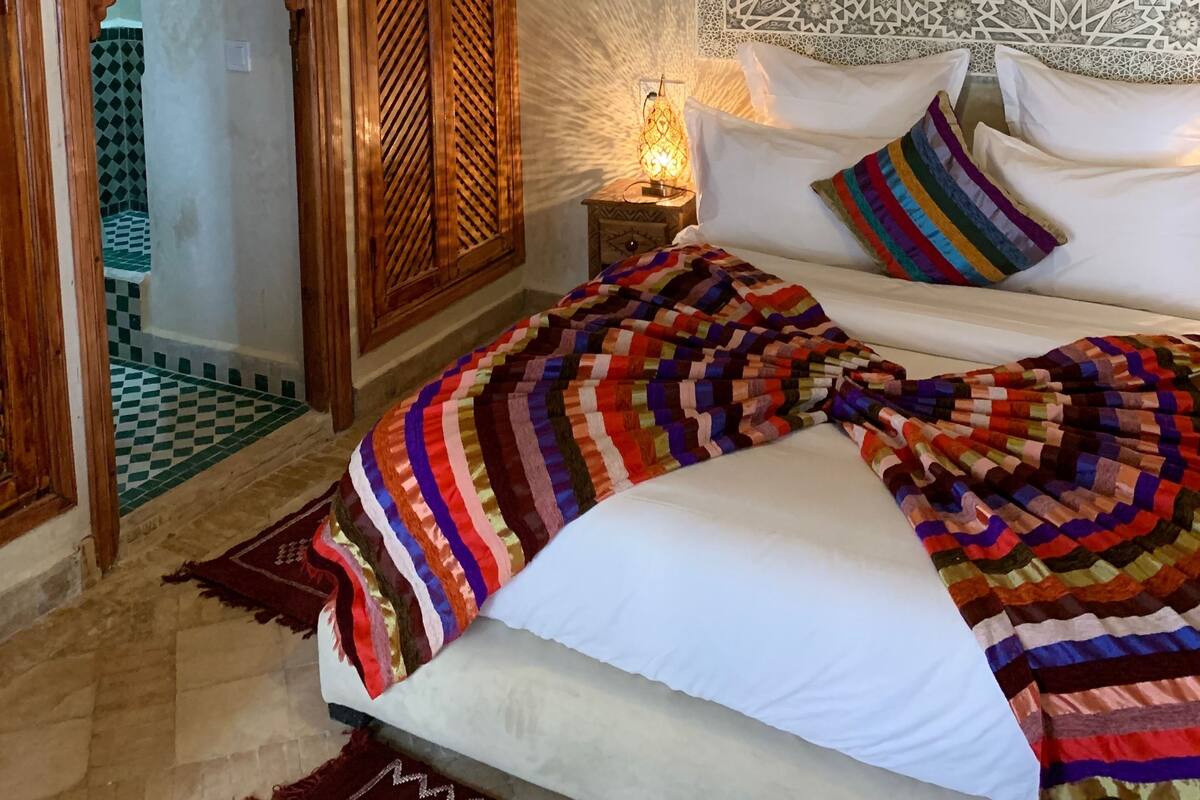 A bedroom with a bed, pillows, a nightstand, a wall tapestry, and an arched doorway.
