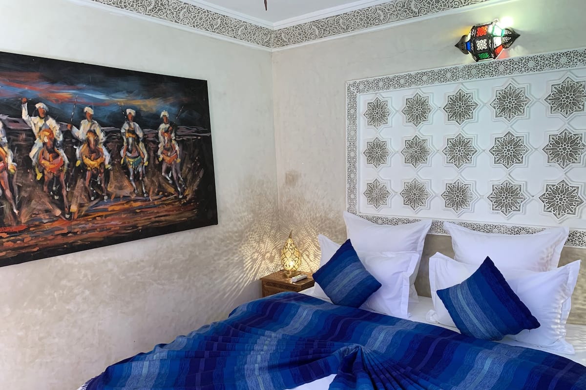 A bedroom with a bed, a painting, a decorative headboard, and a nightstand with a lamp.