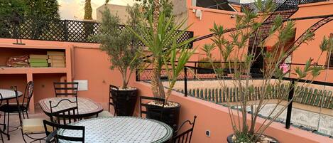 Un patio exterior con mesas y sillas redondas, rodeado de plantas en macetas y una valla decorativa.