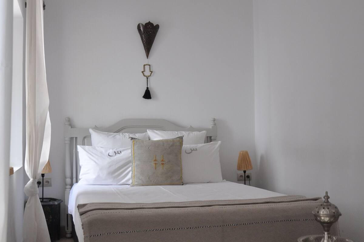 Un dormitorio con cama blanca, una mesita de noche, una lámpara y un objeto decorativo sobre un pedestal verde.