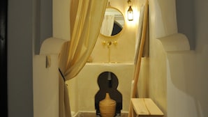 A narrow hallway with an arched doorway, a wooden stool, a small table, and a mirror with a light fixture. - Riad Sin (Marrakech)