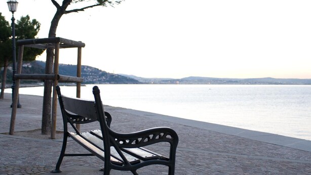 A bench by a waterfront with a stone pathway and a lamp post. - Hotel Coppe (Trieste)