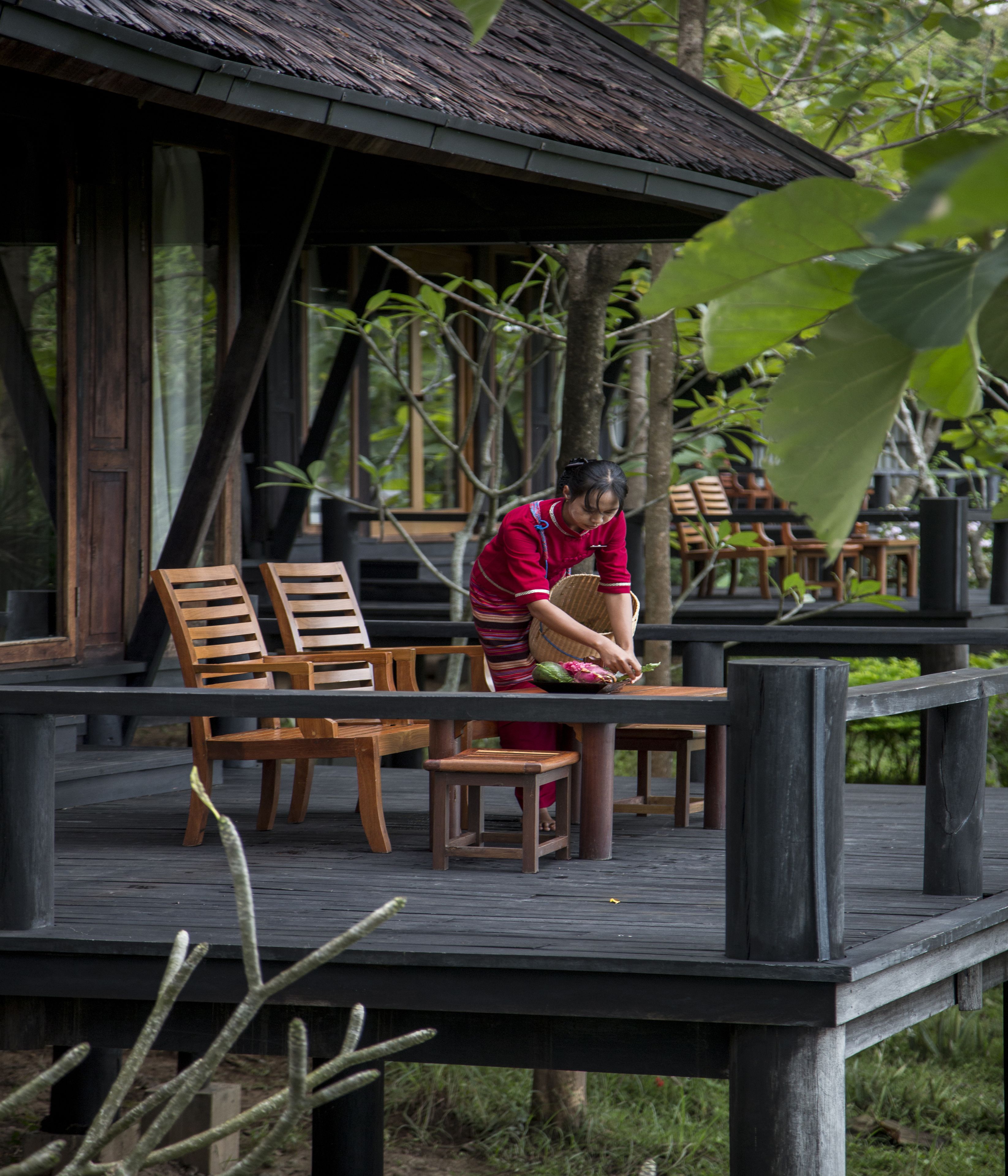 A wooden deck with wooden chairs and tables, a person working with a basket, and a thatched roof structure in the background.