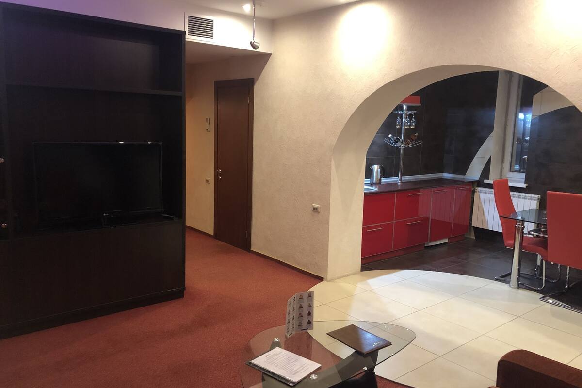 A modern living room with a TV, a glass table, and red furniture. There is a kitchen visible through an archway.