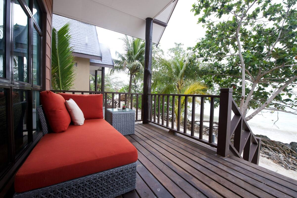 A wooden deck with a red cushioned seating area, a glass balustrade, and a view of the beach and palm trees.