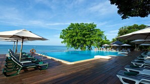 Terraza de madera con sillones de descanso y sombrillas, con vista a una piscina y al mar.