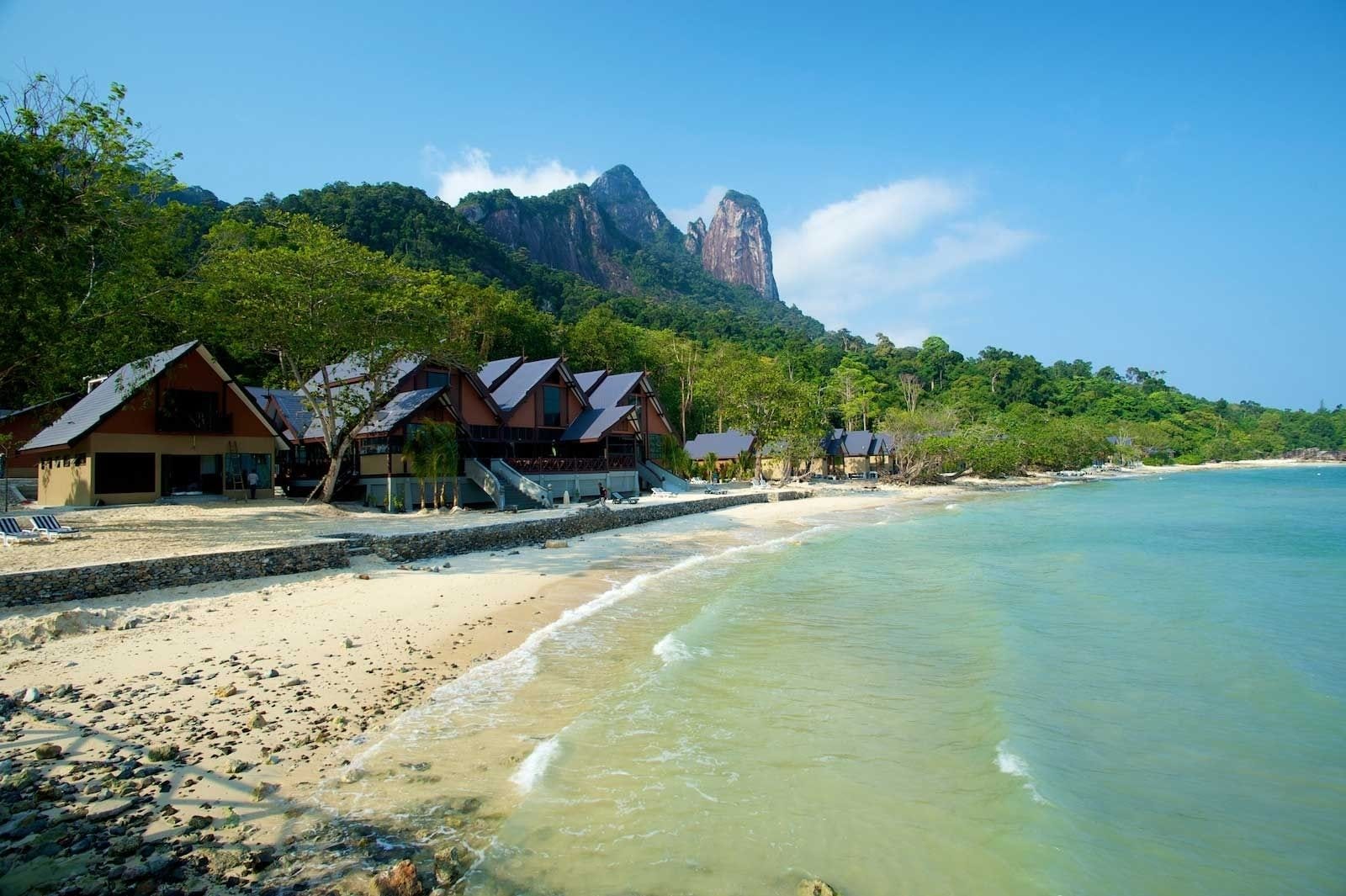 Un resort frente a la playa con varias construcciones tipo bungalow, una playa de arena y montañas al fondo.