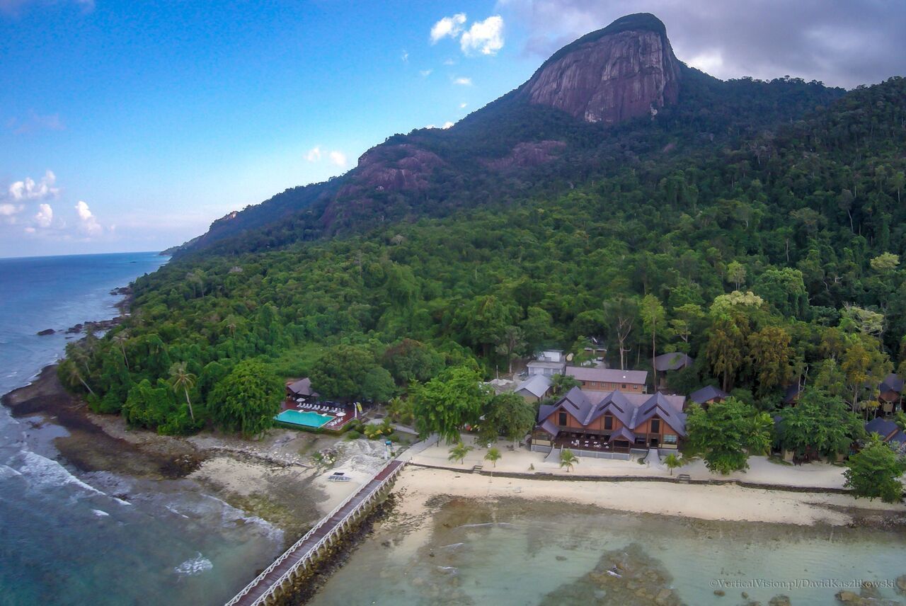 An aerial view of a coastal resort with a prominent mountain in the background, a sandy beach, and several buildings with a traditional architectural style.