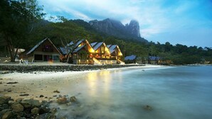 Un hotel frente a la playa con varias edificaciones, un litoral rocoso y una montaña al fondo.