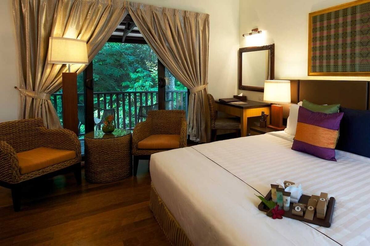 A hotel room with a bed, a desk, a chair, and a view of greenery outside.