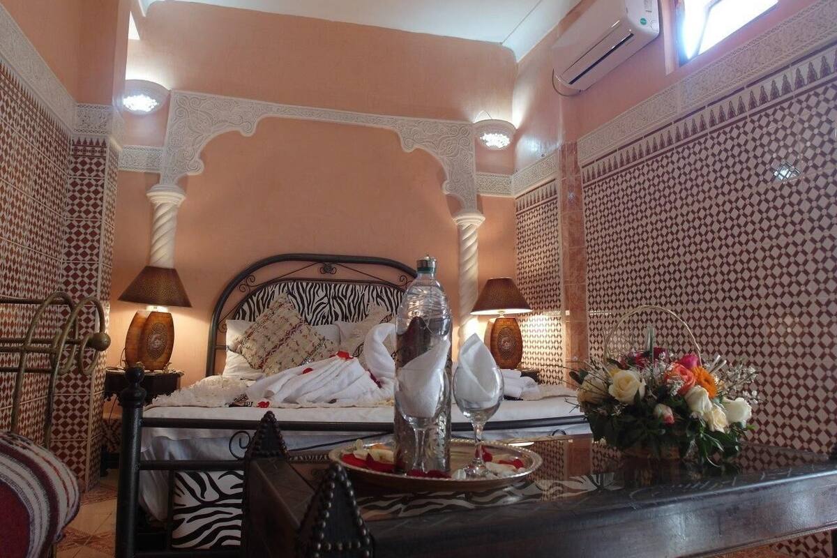 A room with a bed, a bedside table with a lamp, a water bottle, a glass, and a basket of flowers.