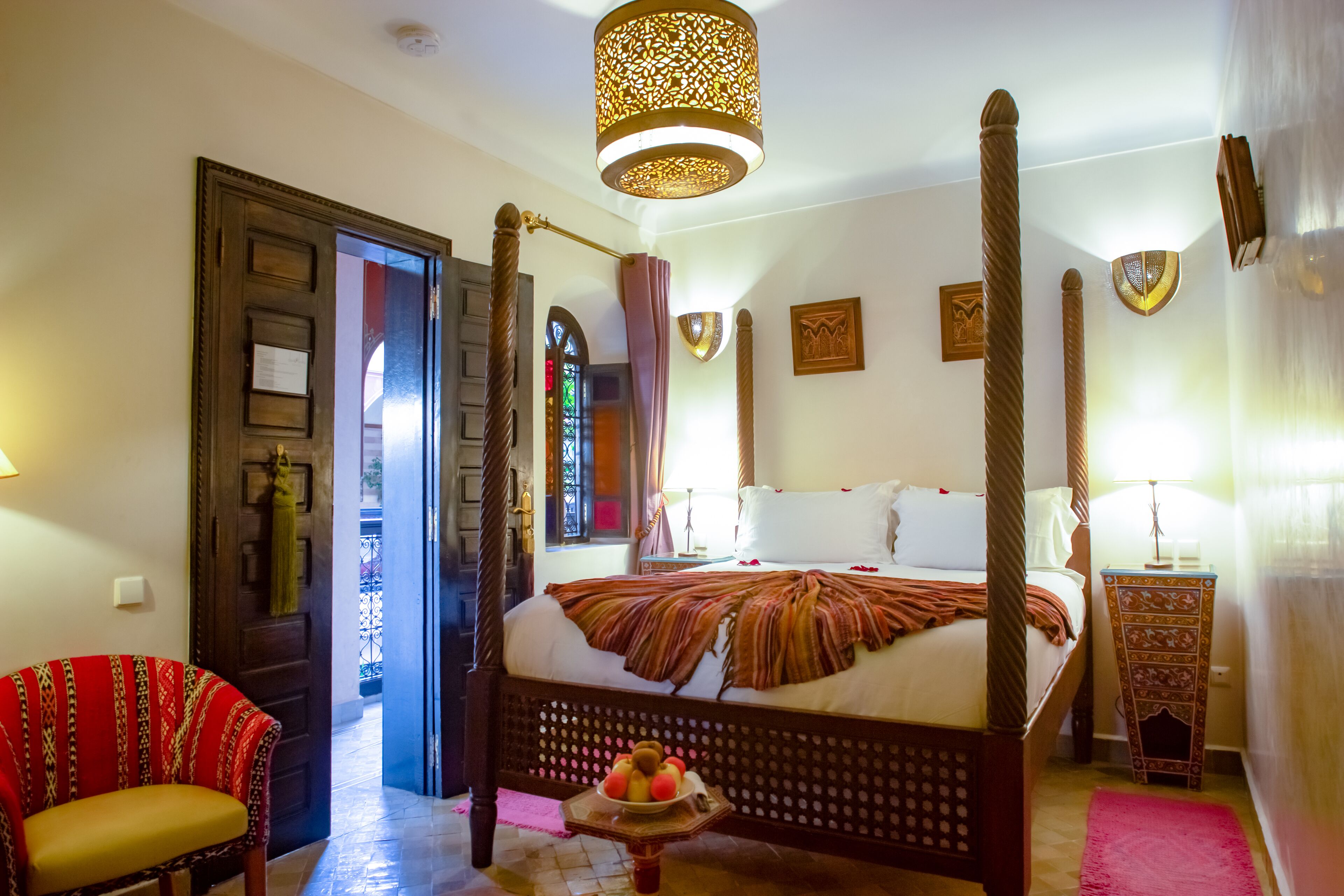 A four-poster bed with a fruit bowl on the footboard, a wooden chair, and a decorative lamp.