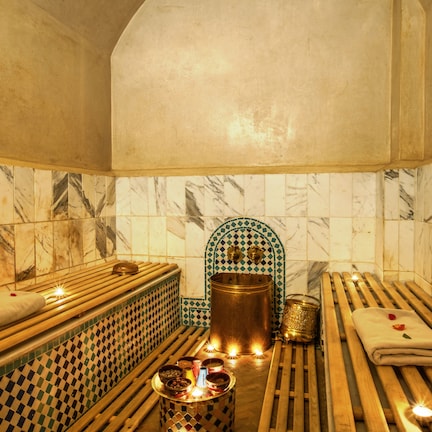 A traditional bath area with wooden benches, a central copper vessel, and a tiled wall with a geometric pattern.