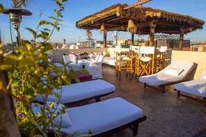 A rooftop lounge area with sun loungers, a thatched umbrella, and a table with chairs.