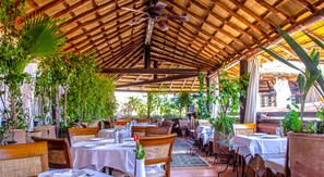 An outdoor restaurant with wooden tables and chairs, surrounded by potted plants and a thatched roof.