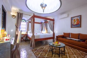 A four-poster bed with a canopy, a sofa, a round table with fruit, and a chandelier.
