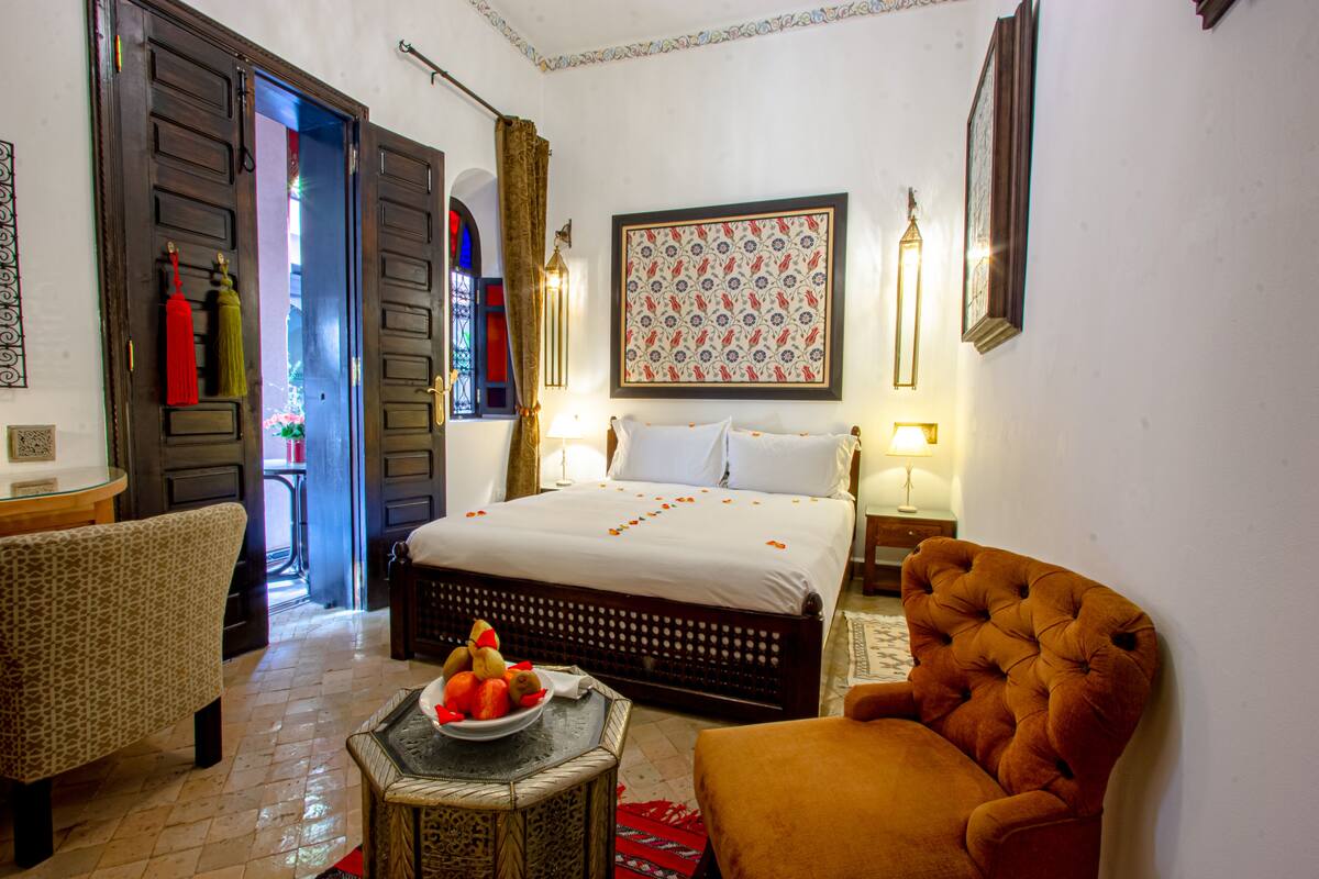 A bedroom with a bed, a chair, a table with fruit, and a wall with a framed pattern.
