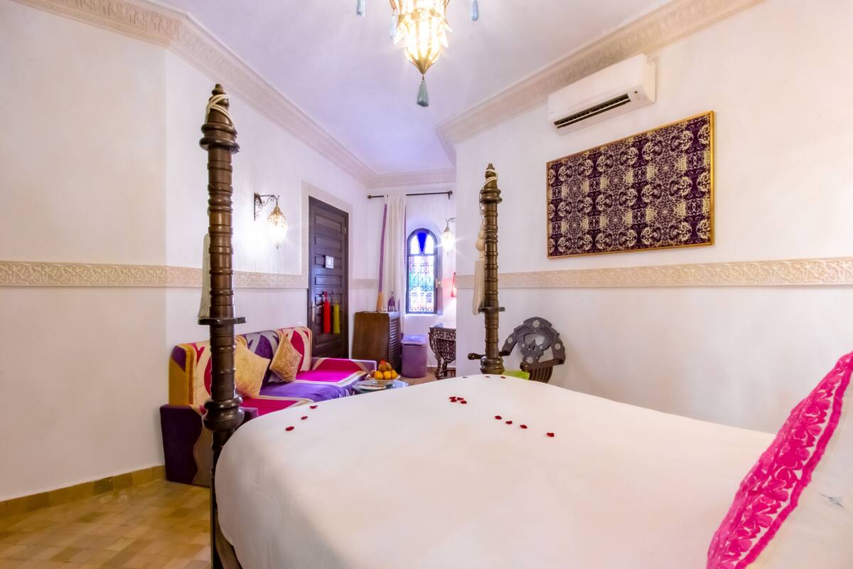 A hotel room with a large bed, a chandelier, a decorative wall hanging, and a view of the outside through a window.