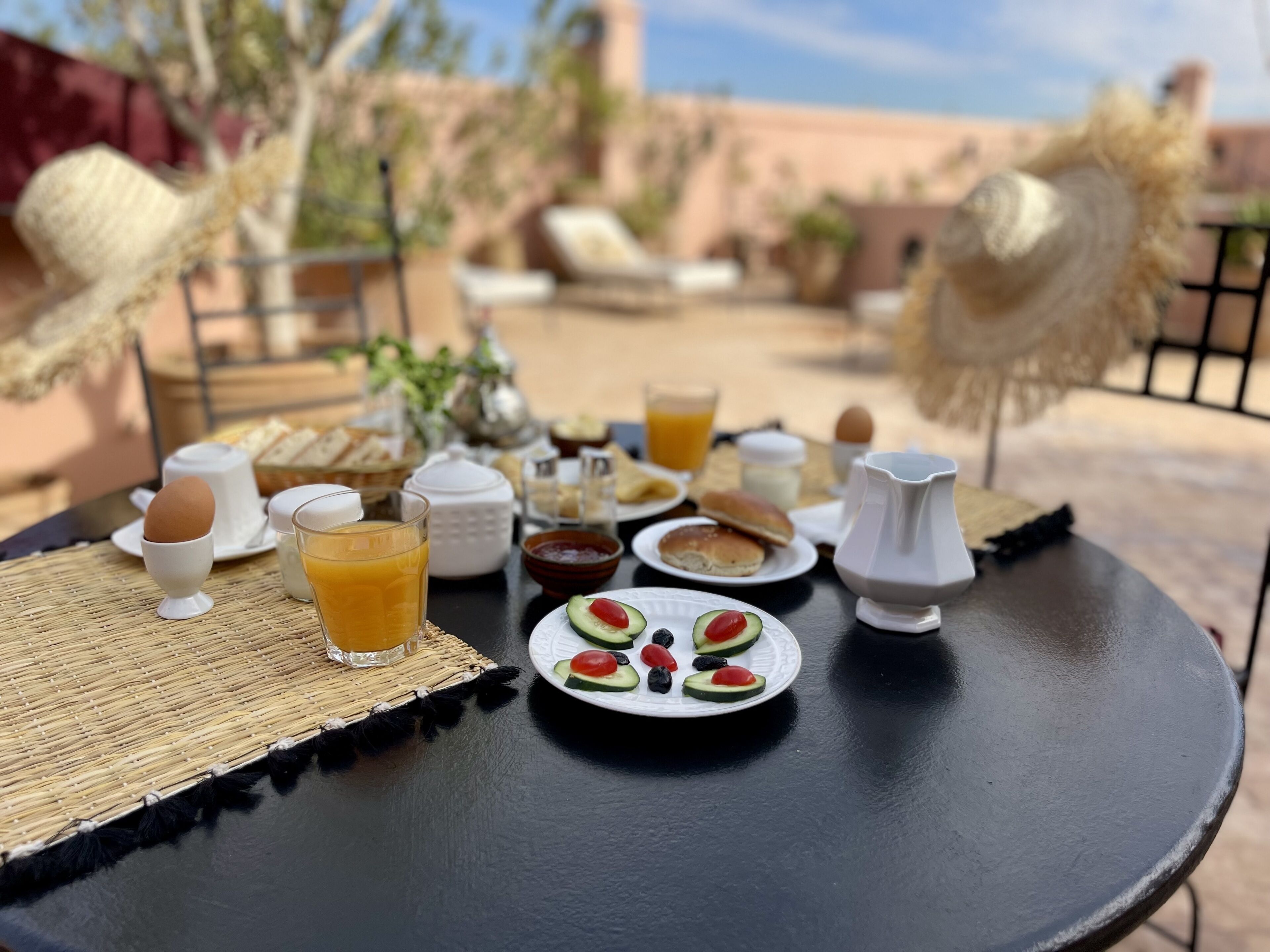 Et morgenbord dækket med forskellige spiser, blandt andet brød, æg og frugt, placeret på en udendørs terrasse med fletmøbler og udsigt over det omkringliggende landskab.