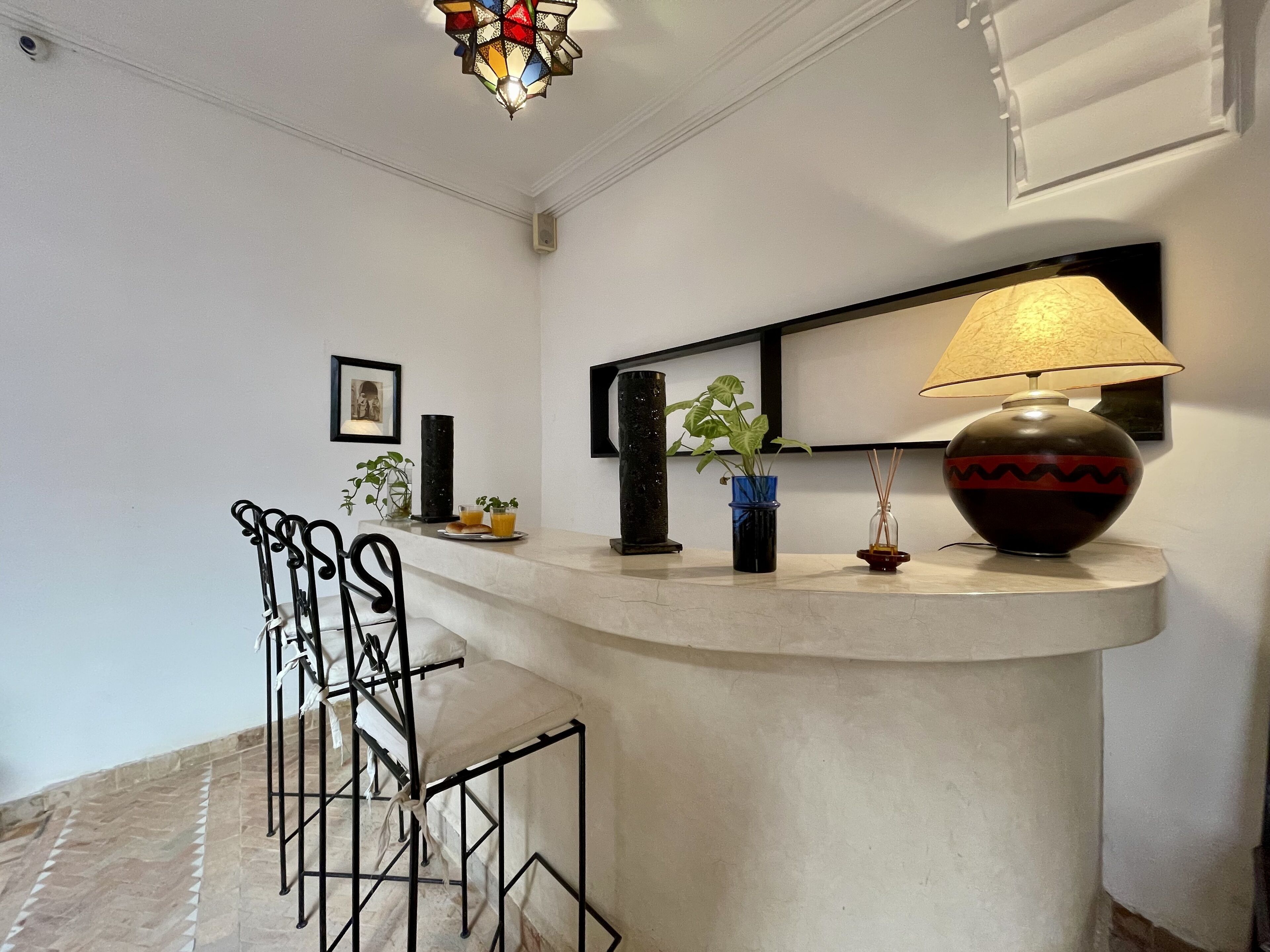 A modern interior with a curved white counter, a black metal chair, a stained glass light fixture, a wall-mounted mirror, and a table lamp.