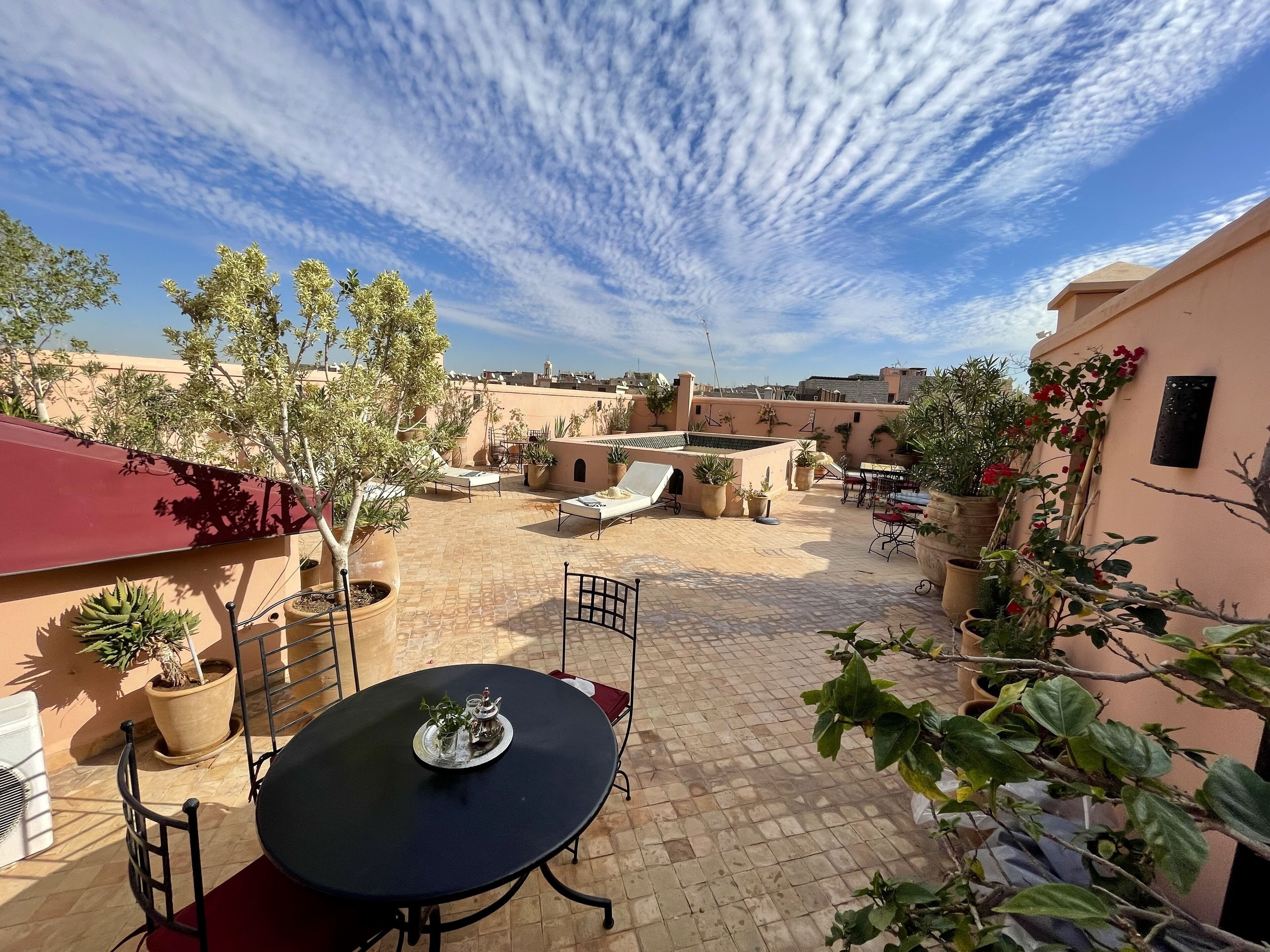 A courtyard with a round table and chairs, surrounded by potted plants and a red awning.