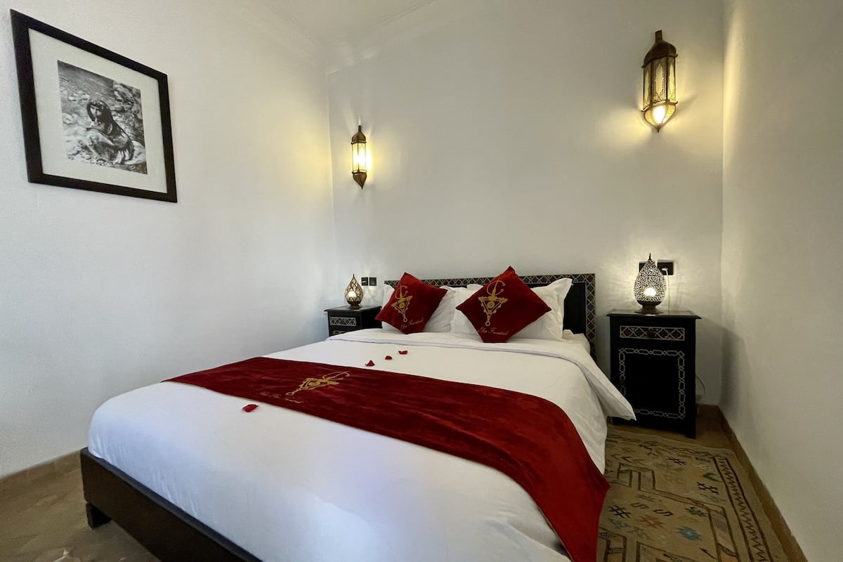A neatly made bed with white linens and a red throw, two bedside lamps, a framed picture on the wall, and a patterned rug on the floor.