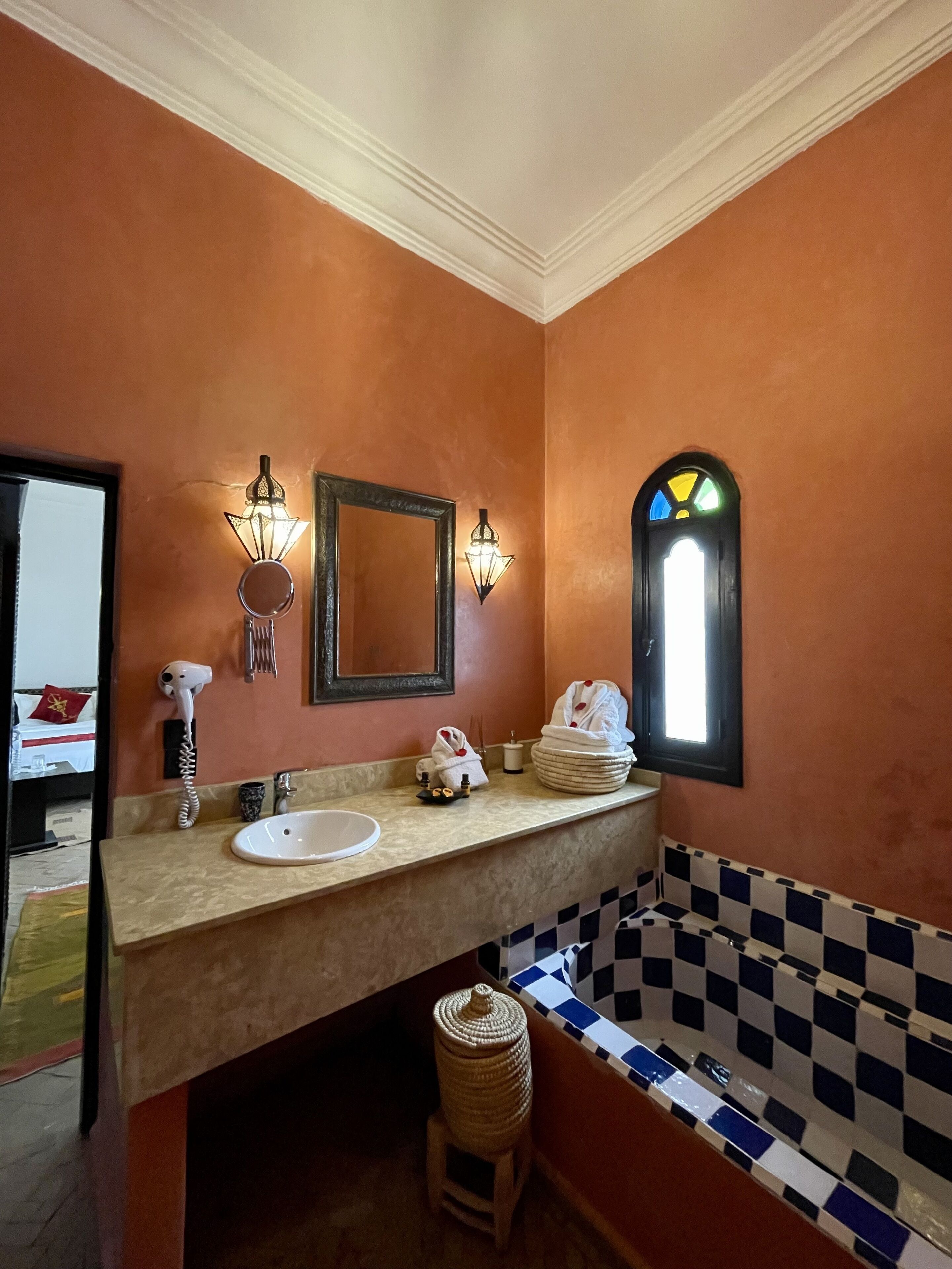 Une salle de bain avec un comptoir beige, un lavabo blanc, un grand miroir, un luminaire fixé au mur, un porte-serviettes et une baignoire à carreaux.