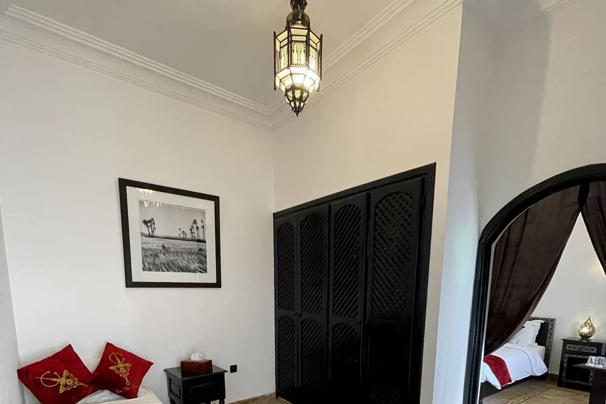 A bedroom with a bed, a nightstand, a framed picture on the wall, a black wardrobe, and a doorway leading to another room.