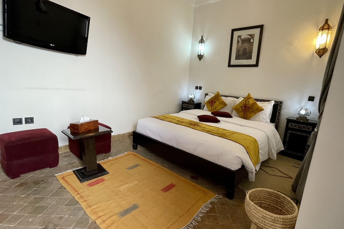 A hotel room with a bed, bedside tables, a TV mounted on the wall, a red ottoman, a patterned rug, and a wall-mounted picture.