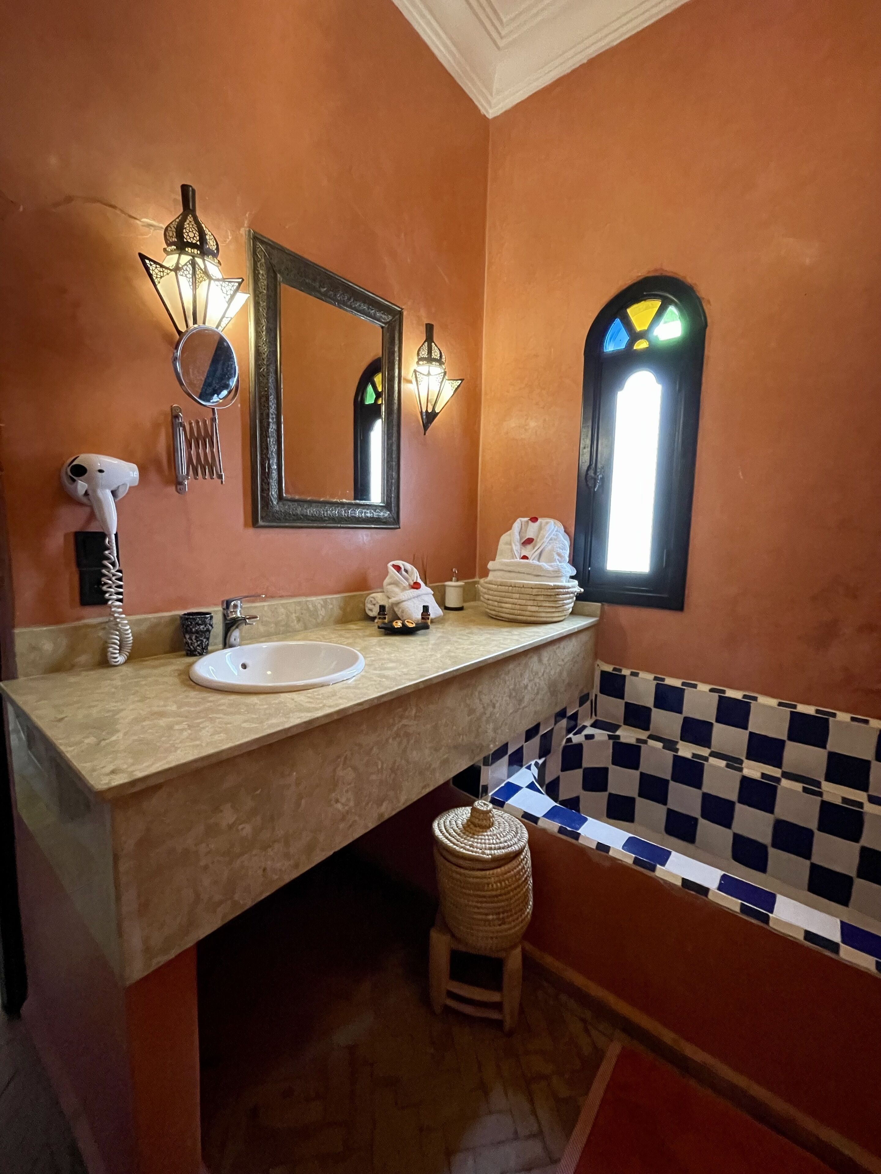 Une salle de bain avec un comptoir beige, un lavabo blanc, un miroir, un luminaire fixé au mur, un porte-serviettes, un sèche-cheveux, un petit tabouret en osier et une baignoire à carreaux.