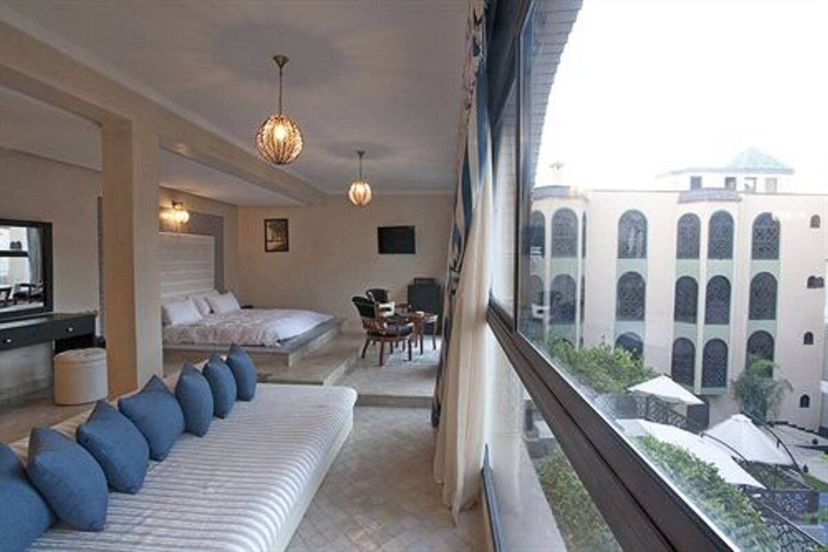 A hotel room with a bed, a sofa, a dining area, and a view of a building with arched windows.