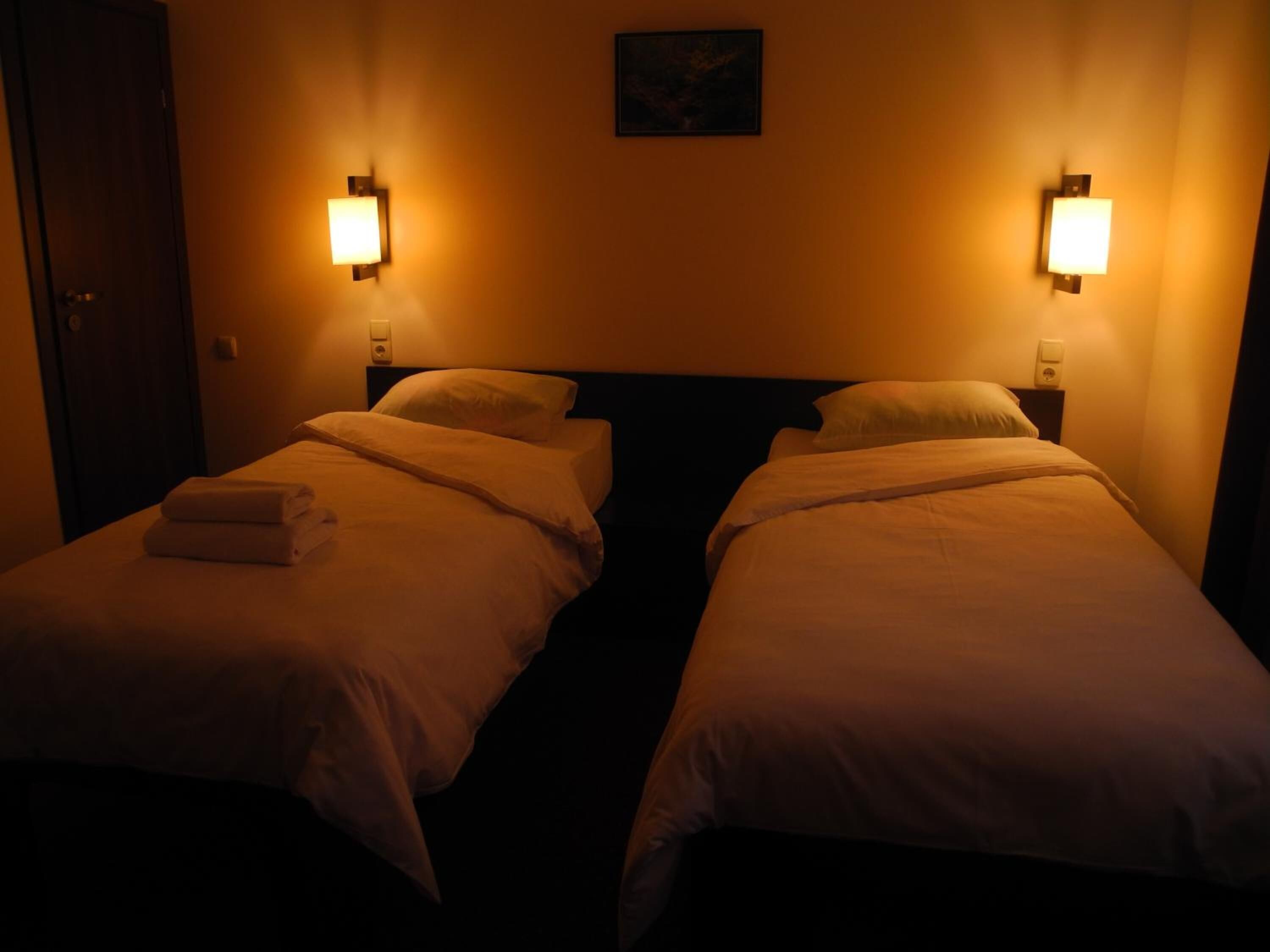 A hotel room with two single beds, each with a nightstand and a wall-mounted lamp.